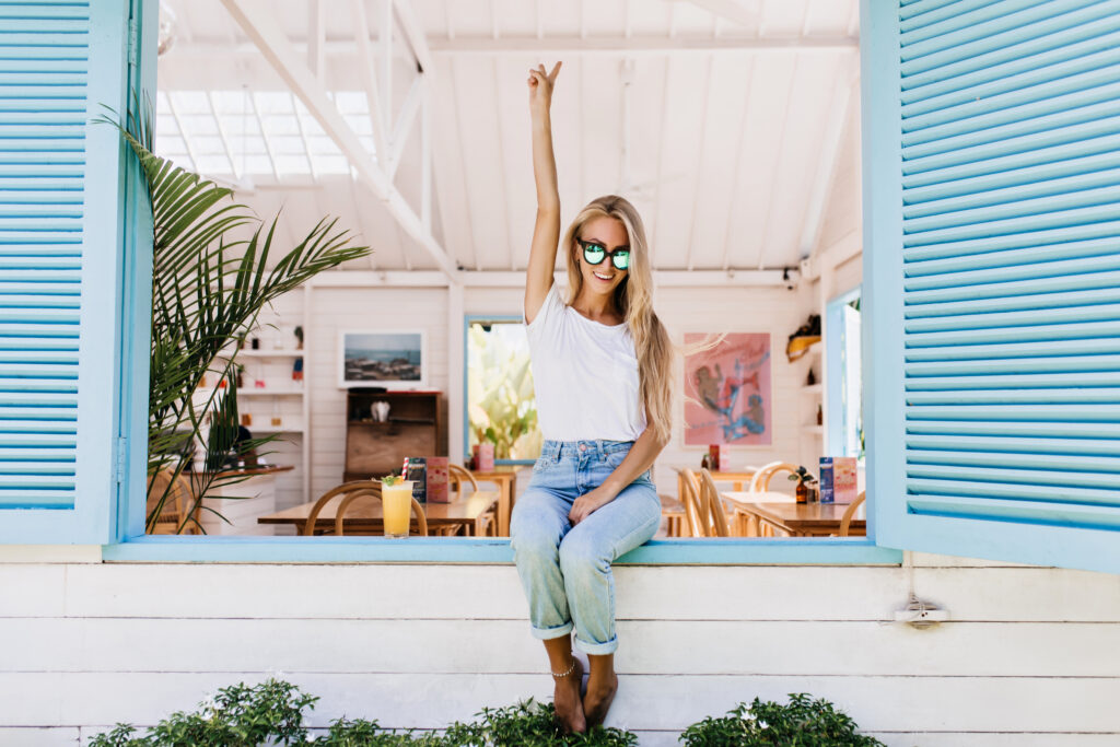 Funny barefooted girl in white t-shirt expressing energy. Photo of gorgeous blonde lady in jeans sitting on window sill with orange cocktail.